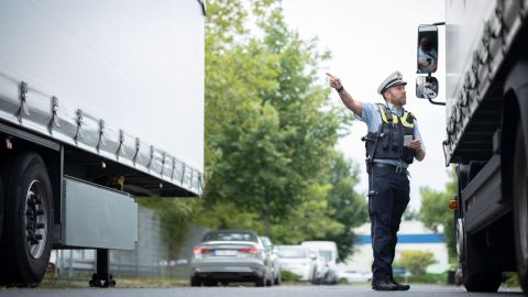 Uniformierter Polizist mit weißer Mütze auf dem Kopf steht auf der Straße zwischen zwei LKWs. Er spricht mit dem Fahrer des rechten LKWs und zeigt dabei mit dem Arm nach links. Im Hintergrund stehen parkende Pkws.