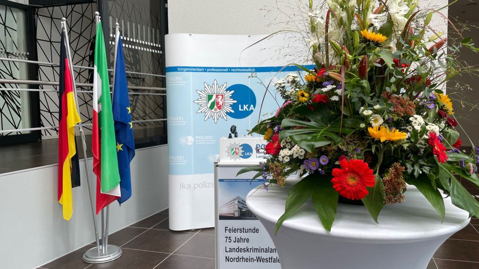 Schild mit Hinweis auf Feierstunde 75jähriges Jubiläum LKA NRW und Blumen auf Stehtisch