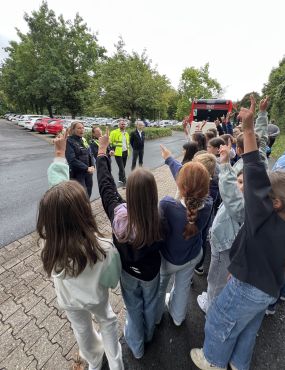 Über den Rücken von Kindern, die sich melden, sind Polizistinnen und Mitarbeiter der Märkischen Verkehrsgesellschaft zu sehen. Im Hintergrund steht ein Linienbus.